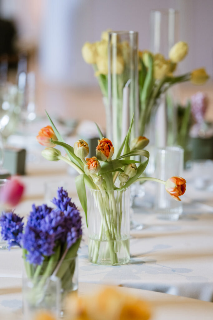 A collection of clear glass bud vases arranged as a centerpiece on a wooden table, featuring vibrant orange tulips or ranunculus and purple hyacinths.