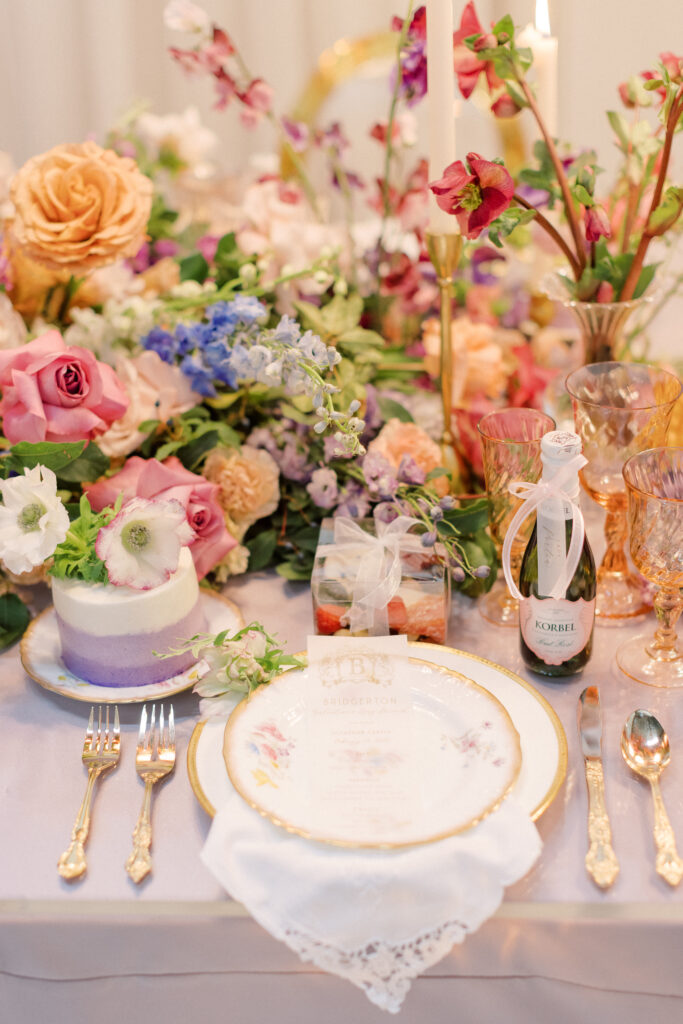 Bridgerton wedding inspiration with a tablescape featuring a pastel color palette of lavender, peach, pink, and blue. Floral arrangements include peonies, garden roses, and anemones. Candles of varying heights illuminate the setting, while linens with intricate patterns and lace enhance the design