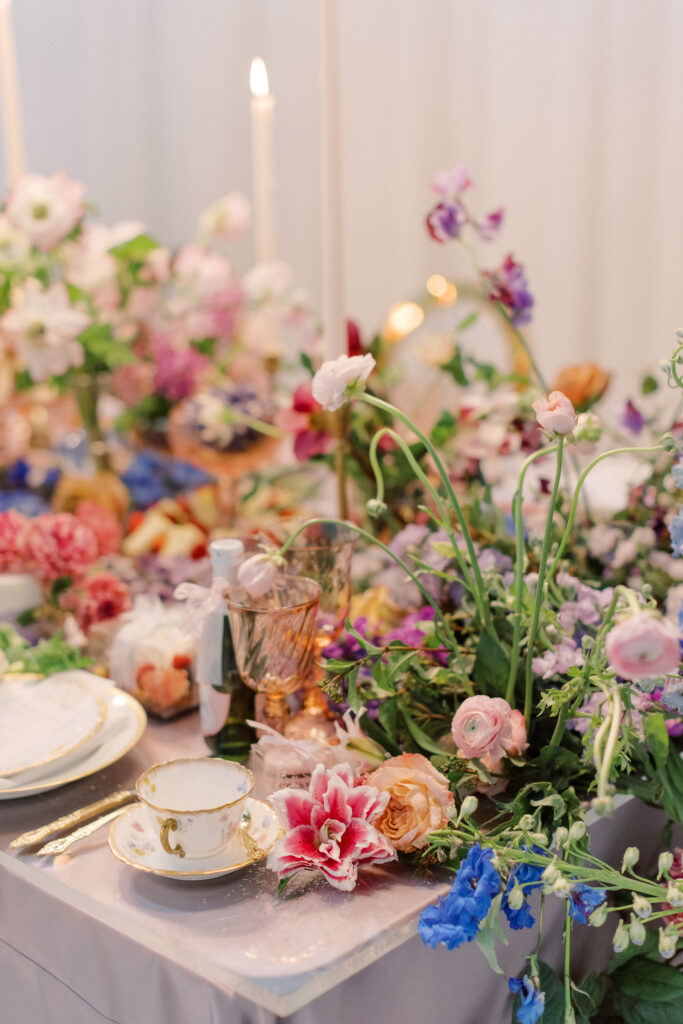 Bridgerton wedding inspiration with a tablescape featuring a pastel color palette of lavender, peach, pink, and blue. Floral arrangements include peonies, garden roses, and anemones. Candles of varying heights illuminate the setting, while linens with intricate patterns and lace enhance the design