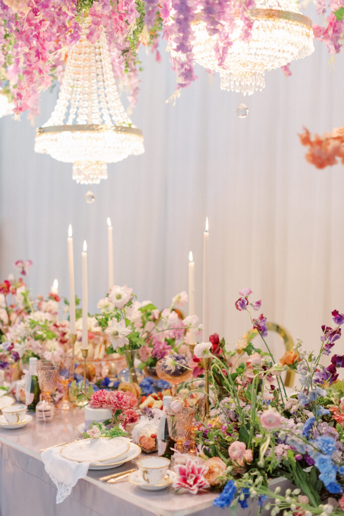 Bridgerton wedding inspiration with a tablescape featuring a pastel color palette of lavender, peach, pink, and blue. Floral arrangements include peonies, garden roses, and anemones. Candles of varying heights illuminate the setting, while linens with intricate patterns and lace enhance the design