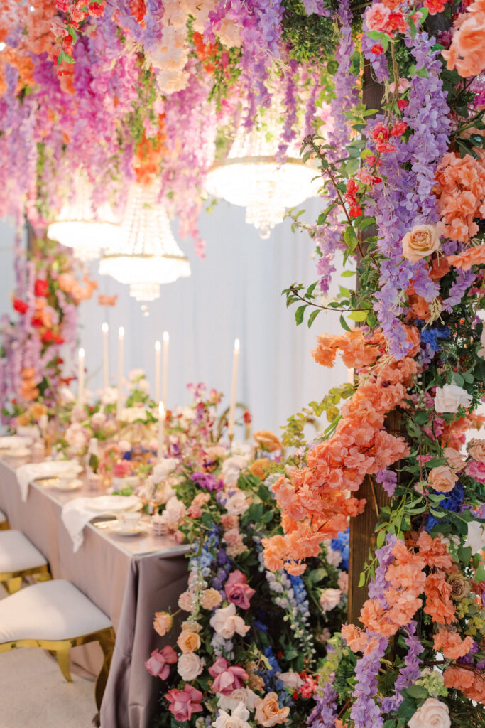 Bridgerton wedding inspiration with a tablescape featuring a pastel color palette of lavender, peach, pink, and blue. Floral arrangements include peonies, garden roses, and anemones. Candles of varying heights illuminate the setting, while linens with intricate patterns and lace enhance the design
