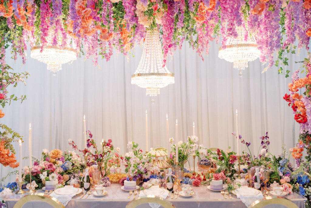 Bridgerton wedding inspiration with a tablescape featuring a pastel color palette of lavender, peach, pink, and blue. Floral arrangements include peonies, garden roses, and anemones. Candles of varying heights illuminate the setting, while linens with intricate patterns and lace enhance the design